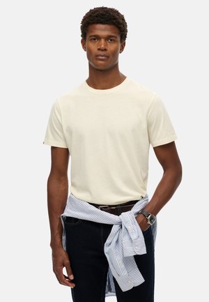 Hellbeige T-Shirt mit kurzen Ärmeln aus weichem Stoff, kombiniert mit dunklen Jeans und einem hellblauen gestreiften Hemd, das um die Taille gebunden ist.