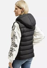 Czarna kamizelka puchowa z kapturem, z poziomym pikowanym wzorem. Noszona na białej bluzie z czarnym abstrakcyjnym haftem na rękawach.