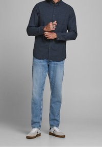 Tummansininen pitkähihainen nappipaita, jossa on rintatasku, yhdistettynä vaaleansinisiin farkkuihin ja valkoisiin lenkkareihin, joissa on ruskeat pohjat.