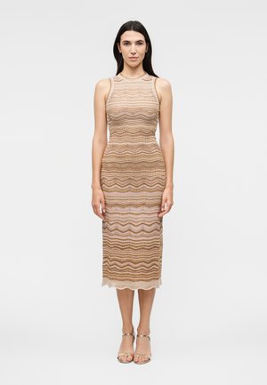 Γυναίκα στέκεται φορώντας ένα αμάνικο, εφαρμοστό, μήκους μέχρι τη μέση της γάμπας φόρεμα με κυματιστό ριγέ μοτίβο σε μπεζ και καφέ αποχρώσεις και χρυσά τακούνια με λουράκι στον αστράγαλο.