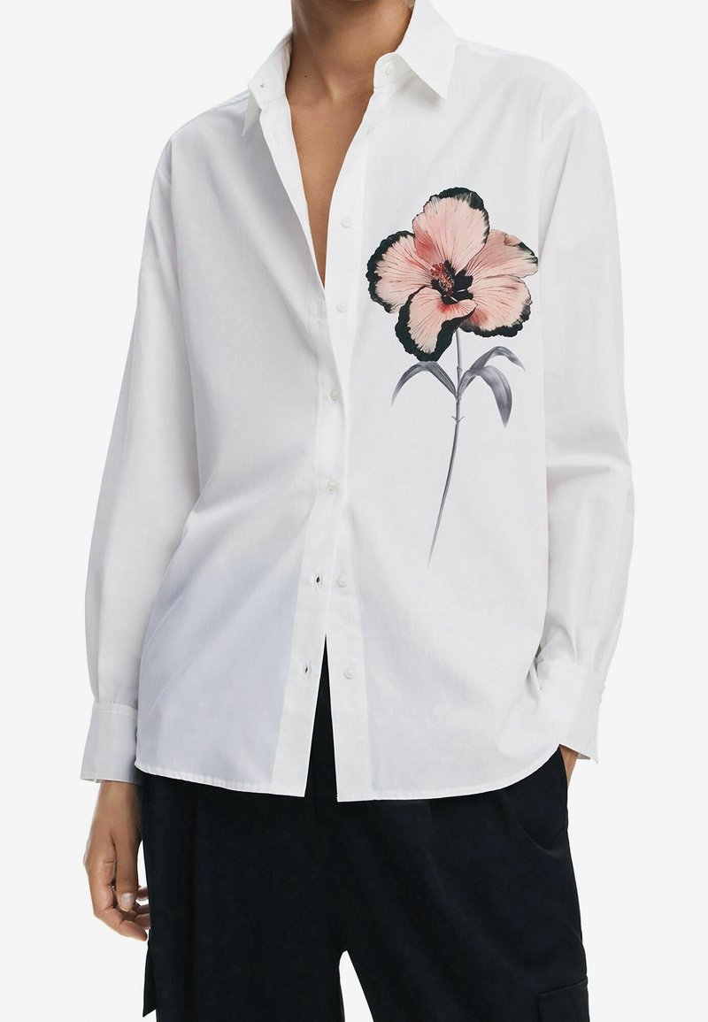 Chemise blanche à manches longues avec boutons, motif de fleur rose et noire sur la poitrine gauche, portée avec un pantalon noir.