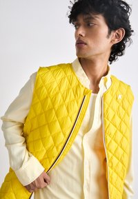 Gilet matelassé jaune avec fermeture éclair et rayure multicolore, porté sur une chemise jaune clair. Présente un motif en losanges et aucun logo visible.
