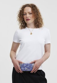 Blue floral-patterned handbag with gold accents held in hands. Model wears a white short-sleeve t-shirt, showcasing a simple design.