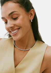 Sølvfarvet choker med funklende rhinsten, parret med matchende sølvfarvede stud øreringe. Båret over et beige V-halsplagg.