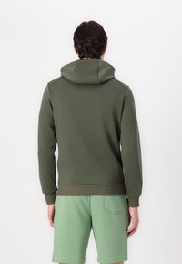 Hoodie u maslinasto zelenoj boji, s opuštenim krojem, rebrastim donjim dijelom i rukavima, u kombinaciji s svijetlozelenim kratkim hlačama s stražnjim džepom.