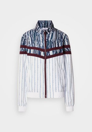 Hvid jakke med blå lodrette striber og abstrakte mønstre. Har mørkerøde bånd over brystet, høj krave og elastiske manchetter.