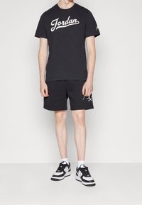 Osoba oblečená v černém tričku Jordan a odpovídajících černých šortkách s bílými a černými teniskami Nike na jednobarevném pozadí.