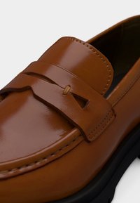 Mocassin en cuir marron à finition polie, avec une lanière décorative sur le dessus et une semelle noire visible sur le bord inférieur.