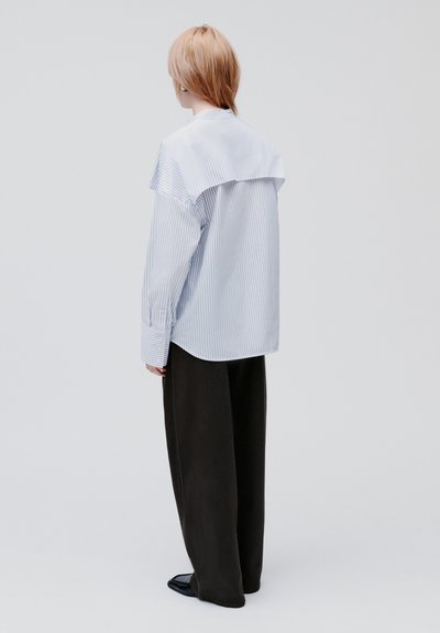 Gestreiftes Hemd in Hellblau und Weiß mit einer großen, geschichteten Rückenpasse, kombiniert mit weiten schwarzen Hosen, minimalistischem Design.
