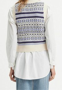 Gilet en tricot crème avec des motifs géométriques bleus, navy et blancs. Il présente un ourlet côtelé et un design sans manches, superposé sur une chemise blanche à boutons.