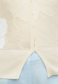 Gros plan d’un cardigan tricoté crème avec fermeture à boutons et motif floral en maille ajourée sur tissu bleu clair.