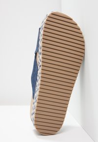 Sula av en beige och blå espadrille-sandal med en tjock, ribbad gummisula och vävt repkant mot en vit bakgrund.