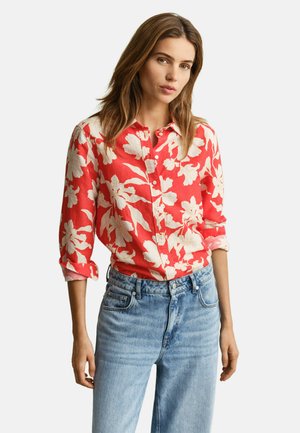 Femme portant une chemise rouge à boutons avec un grand imprimé floral blanc et des manches retroussées, assortie à un jean taille haute bleu clair.