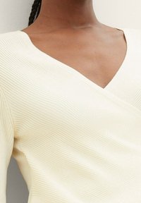 Gräddfärgad ribbad v-ringad topp med en figurnära siluett och texturerad tyg, som visar en djup halsringning och subtila sömdetaljer.