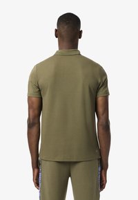 Polo shirt in verde oliva con una trama uniforme, maniche corte e colletto classico. Presenta un orlo dritto e un branding minimale nella parte inferiore.