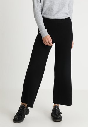 Osoba ubrana w czarne spodnie o szerokich nogawkach, czarne trampki oraz jasnoszary sweter z długim rękawem, stojąca na tle jednolitego białego tła.