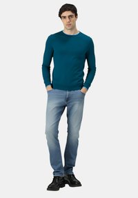 Giovane uomo che indossa una camicia a maniche lunghe color turchese, jeans azzurri chiari e scarpe eleganti nere, in piedi con le mani in tasca su sfondo bianco.