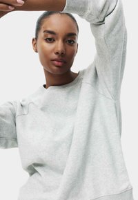 Junge Frau mit glatter Haut und nach hinten getragenem Haar, die einen hellgrauen Sweatshirt trägt und ihren Arm über ihren Kopf hebt, vor einem schlichten Hintergrund.