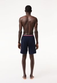 Costumi da bagno blu navy con una cintura elasticizzata che presenta strisce rosse e bianche. Il materiale sembra leggero; i pantaloni arrivano sopra il ginocchio.