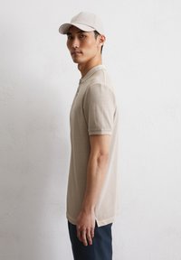 Beżowa koszulka polo z kołnierzykiem i krótkimi rękawami, z bocznym rozcięciem u dołu. Nosi ją w zestawieniu z dopasowaną jasną czapką.