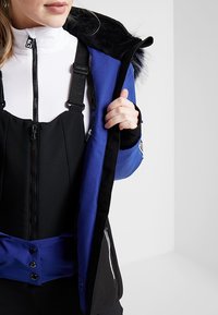 Personne portant une salopette de neige noire par-dessus un haut blanc à fermeture éclair, tenant ouverte une veste d'hiver bleue et noire avec une capuche bordée de fourrure.