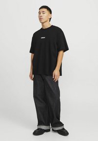 Sort oversized T-shirt med hvidt logo på brystet, parret med løse mørke denimjeans og sorte sko med en kontrastfarvet kant.