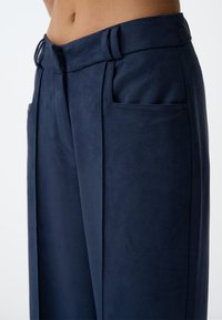 Donkerblauwe broek van een zachte, suede-achtige stof met een hoge taille, riemlussen en zijzakken. Heeft schone, op maat gemaakte lijnen.
