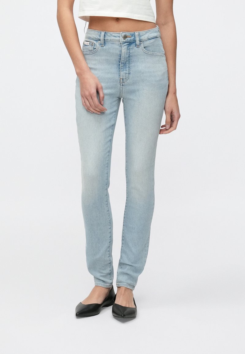 Világoskék, magas derekú, szűk szabású farmer, öt zsebbel és cipzáras sliccel, fekete lapos cipővel párosítva.