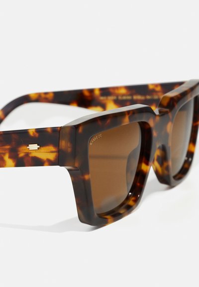 Lunettes de soleil écaille de tortue marron avec de larges montures carrées, verres marron foncé et subtils détails en or sur les branches.