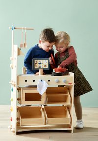 Houten speelkeuken met een lichte kleur, meerdere opbergcompartimenten, hangende keukengerei, blauwe en rode accenten, en een stoffen keukenhanddoek.