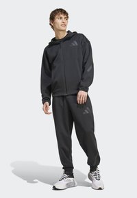 Hoodie preto com fecho, com bolsos à frente, combinado com calças de algodão pretas. Apresenta pequenos detalhes de logótipos e textura. Ténis atléticos brancos.