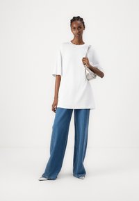 Bijela prevelika majica s kratkim rukavima, u kombinaciji s širokim plavim trapericama i malom metalik srebrnom torbicom. Glatke teksture.