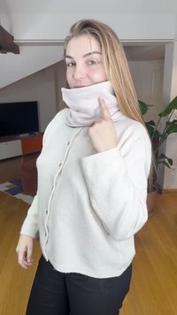 Šviesiai rožinis vilnonis kaklo šildytuvas su subtiliu pilku logotipu, dėvimas po kreminiu džemperiu, minkšta tekstūra ir prigludusiu dizainu. Vidaus aplinka.