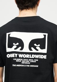 Μαύρο T-shirt με λευκό γραφικό δύο στυλιζαρισμένων προσώπων και κείμενο που απαριθμεί πόλεις και φράσεις «OBEY WORLDWIDE» και «THE MEDIUM IS THE MESSAGE.»