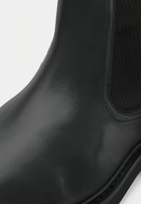 Bota de tornozelo em pele preta com acabamento brilhante, com painéis laterais em malha elástica e sola texturizada. Superfície lisa com costuras limpas.