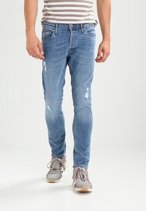 Mand iført lyseblå slidte slim-fit jeans og grå sneakers, der går mod en ensfarvet hvid baggrund.
