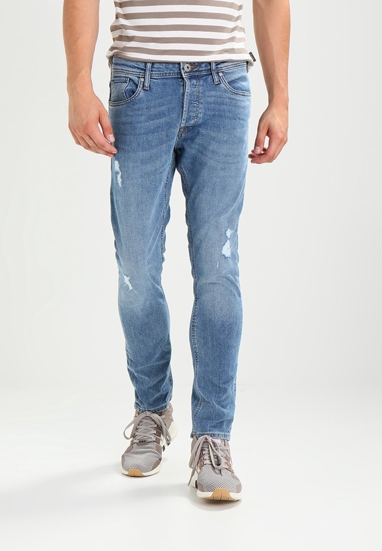 Homme portant un jean slim bleu clair usé et des baskets grises marchant devant un fond blanc uni.