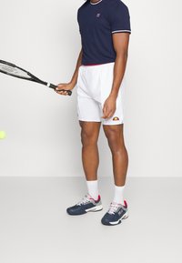 Laivastonsininen urheilupaita valkoisilla reunoilla, valkoiset shortsit logolla ja siniharmaat tenniskengät punaisilla yksityiskohtilla. Pitelee tennismailaa.