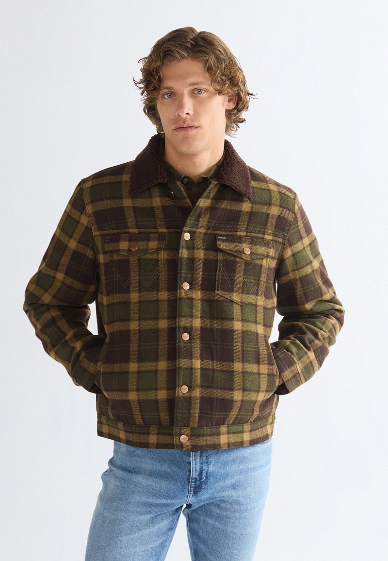 Homme aux cheveux bouclés portant une veste à carreaux verte et marron avec un col marron et un jean bleu clair, mains dans les poches.