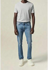 Homme portant un t-shirt blanc uni à manches courtes, un jean droit bleu clair et des baskets blanches, debout devant un fond neutre.