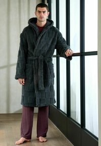 Grå texturerad rock med huva och bälte, som har ett rutmönster, bärs över maroonrandiga byxor, stående vid ett glasfönster.