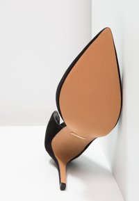 Escarpin en daim noir à talon haut avec un bout pointu, doté d'une semelle texturée marron clair et d'une découpe latérale. Détail accent en or près du talon.