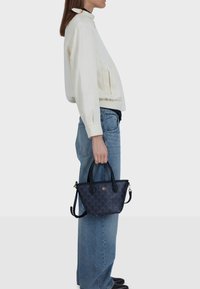 Piccola borsa nera con motivo testurizzato, due manici superiori e tracolla removibile, tenuta da una persona con giacca bianca e jeans.