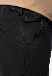 Vaqueros denim negros con corte ajustado, con un bolsillo frontal, costuras sutiles y una textura suave, delineados contra una blusa beige.