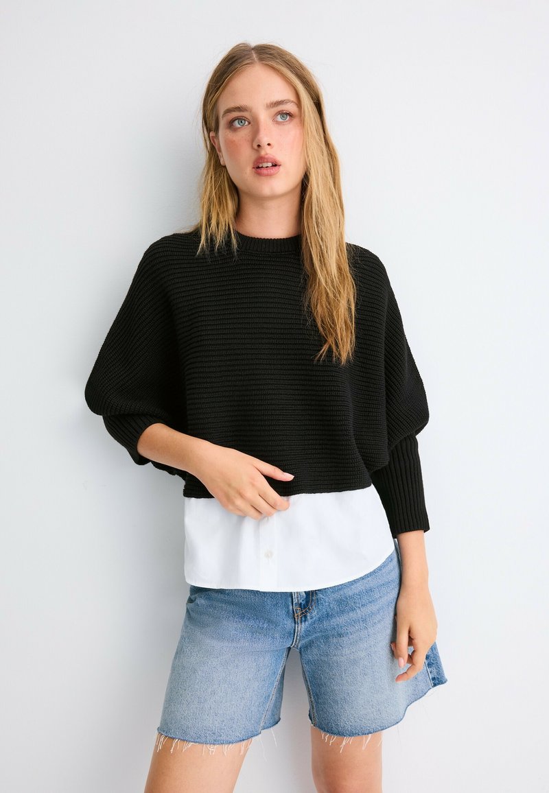 Jeune femme aux longs cheveux portant un pull en maille noir par-dessus une chemise blanche et un short en jean, debout devant un fond blanc.