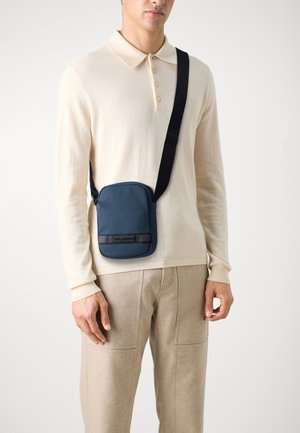 Homme portant un polo beige à manches longues et un pantalon, portant un petit sac bleu en bandoulière avec une sangle noire.