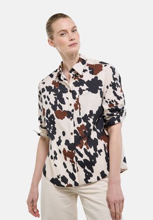 Femme aux cheveux lissés en arrière portant une chemise beige à boutons avec des taches abstraites noires et brunes, associée à un pantalon de couleur claire.