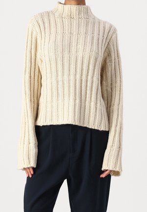 Person wearing a cream ribbed knit sweater with long sleeves and a high neck, paired with loose-fitting black pants.