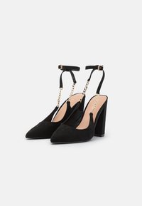 Talons aiguilles en suédine noire avec un talon block, une bride de cheville et un détail en chaîne dorée. Doublure intérieure lisse et fermeture à boucle ajustable.
