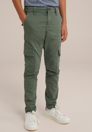 Dziecko ubrane w oliwkowozielone spodnie bojówki z bocznymi kieszeniami i białe trampki, stojące z rękami w kieszeniach.